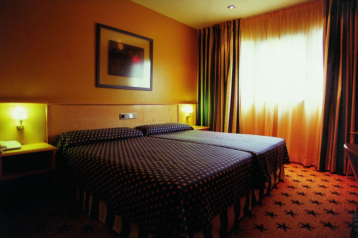 Quarto de hotel com duas camas, uma mesa de cabeceira, uma luminária, um quadro na parede e uma janela com cortinas.