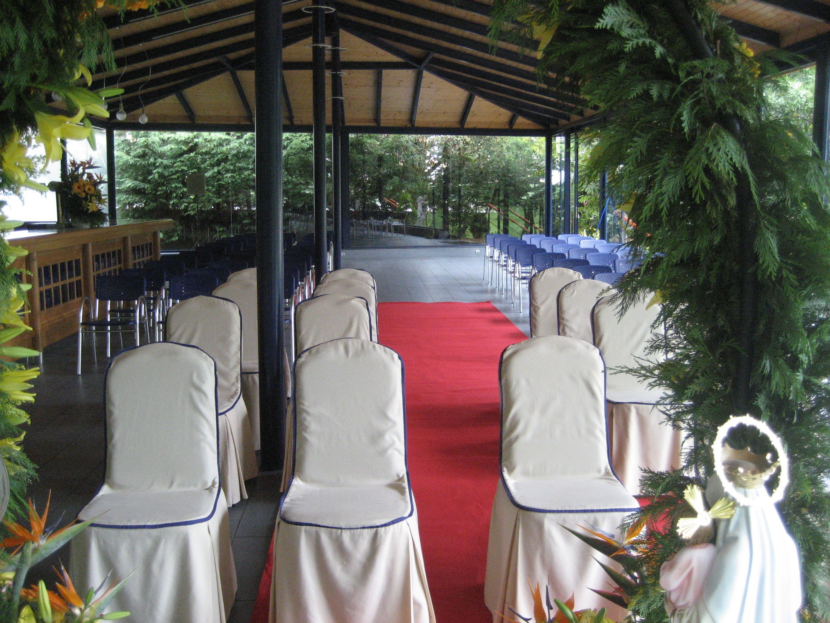 A covered outdoor area with rows of chairs and a red carpet, surrounded by greenery.