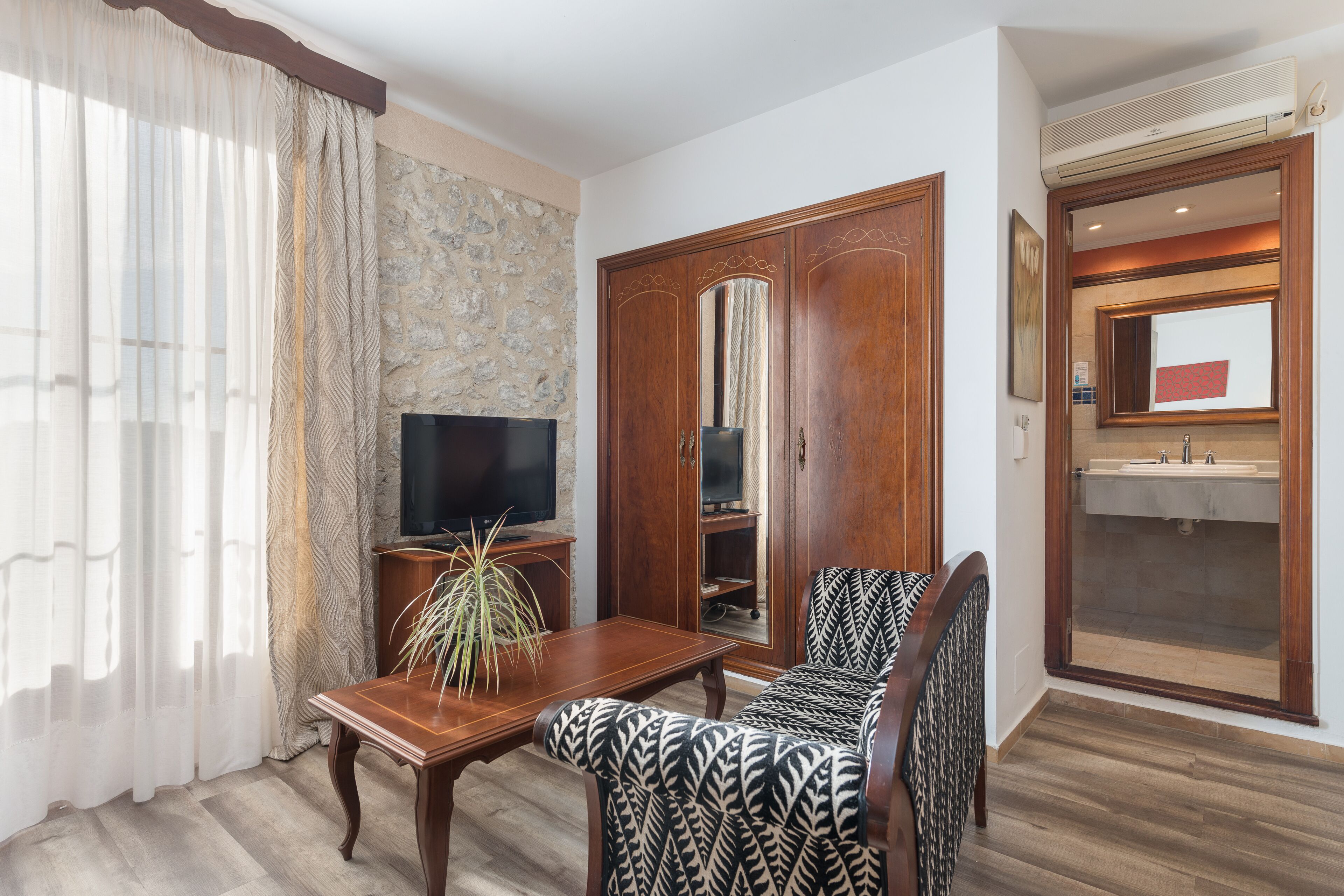 A living room with a stone wall, wooden furniture, a television, and a bathroom with a sink and mirror.