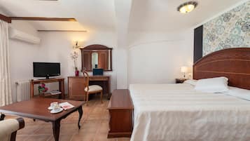 Habitación de hotel con una cama, un escritorio con televisor, una silla, una mesita con una flor y una mesita de noche con lámpara.
