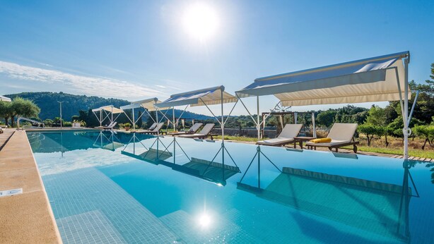 A swimming pool with sun loungers and umbrellas, surrounded by a landscaped area and a view of hills.