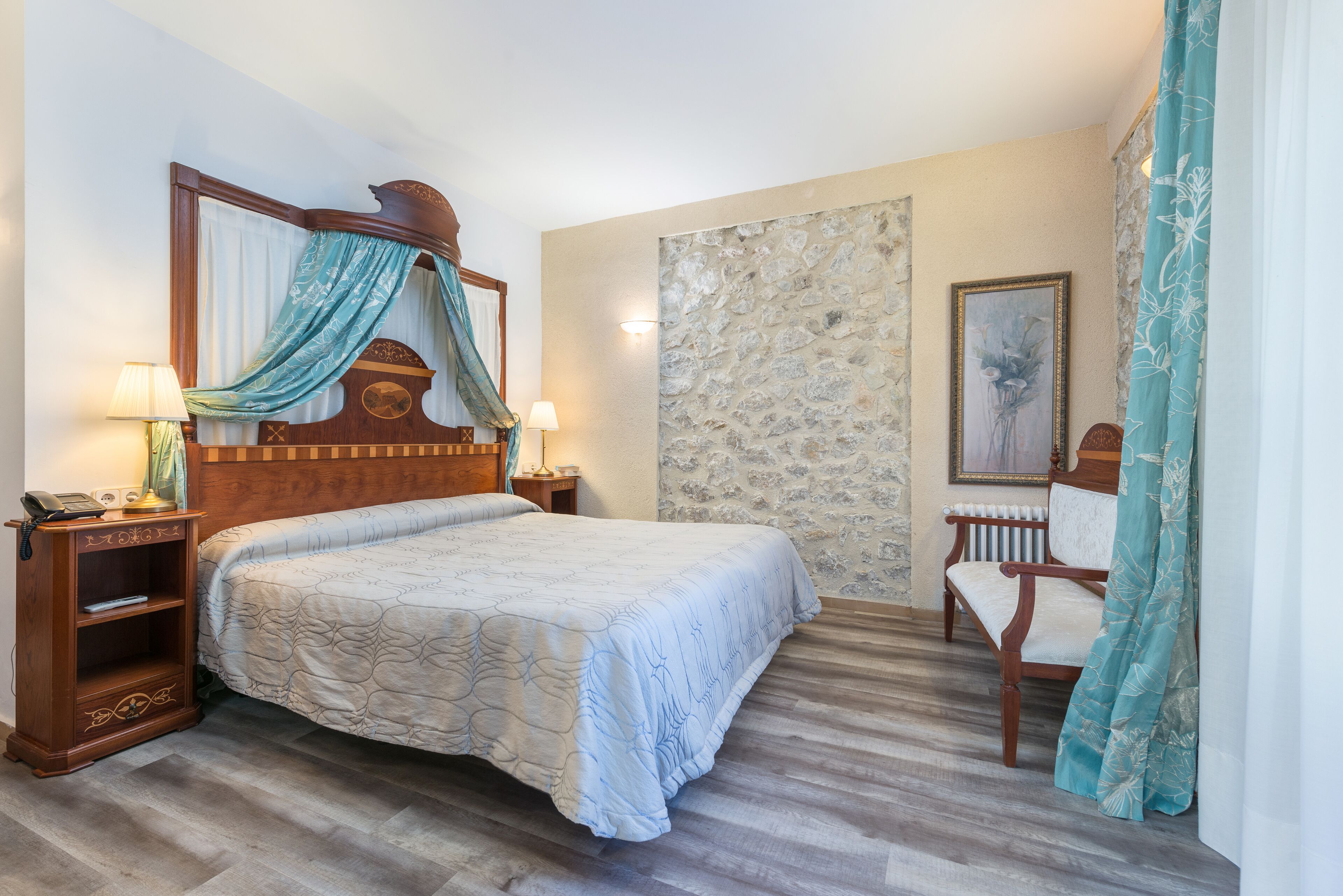 Un dormitorio con una cama grande, cabecera de madera, pared de piedra, una silla y una mesita de noche con lámpara.