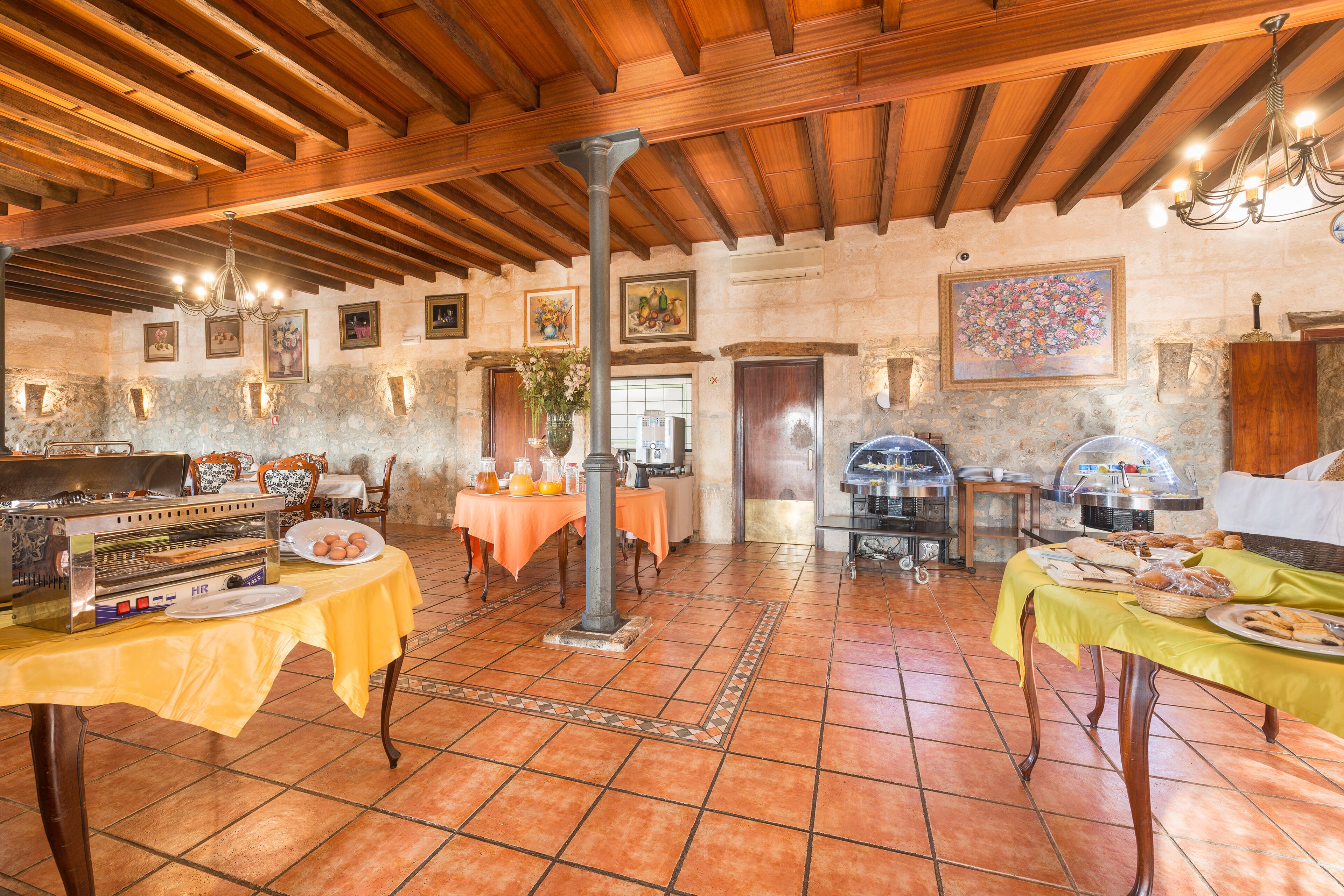 Uma área de refeições rústica com vigas de madeira, paredes de pedra e piso ladrilhado. Há mesas com toalhas amarelas, um espaço para buffet e um candelabro.