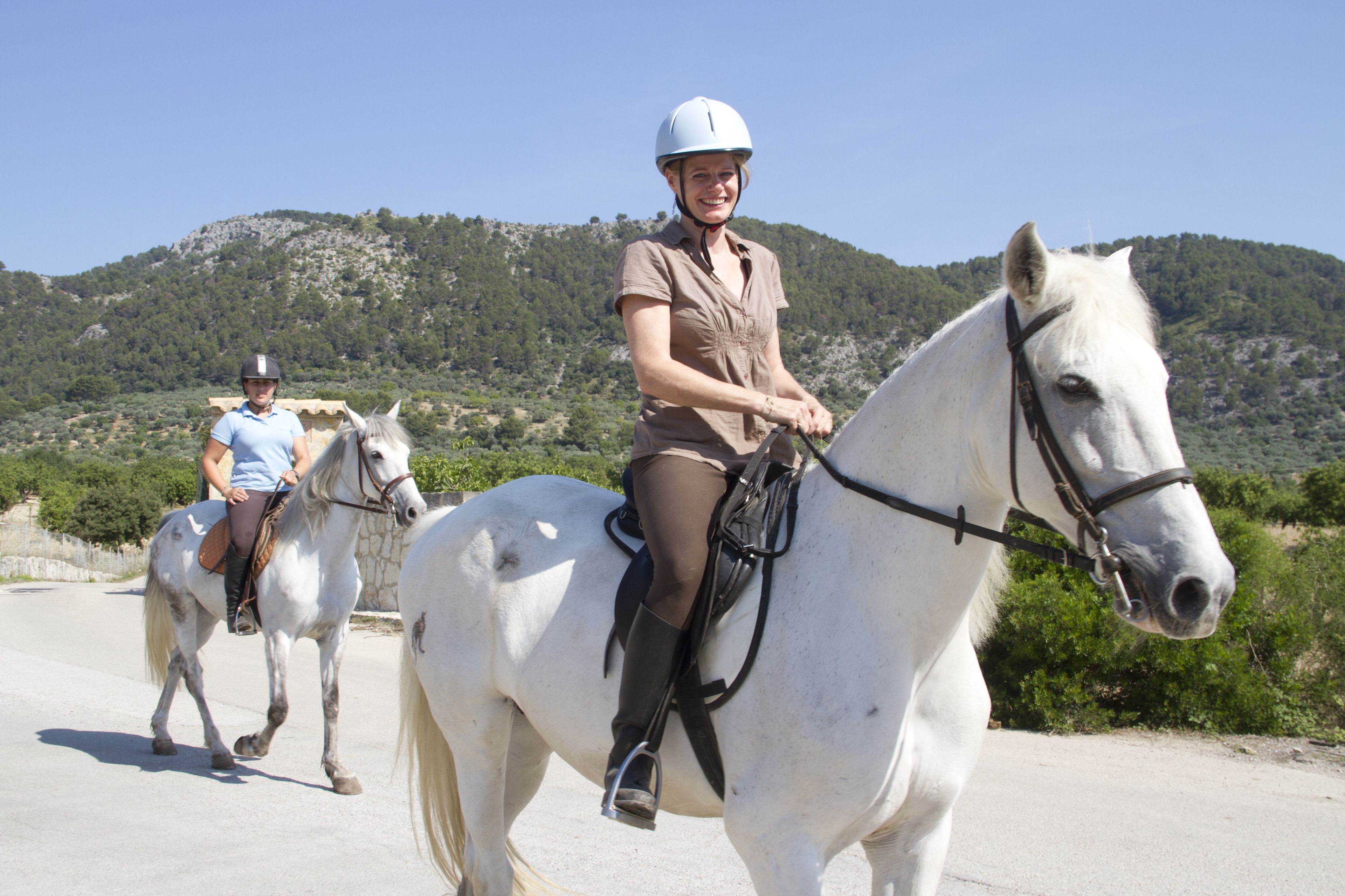 Deux personnes montent des chevaux blancs sur un chemin pavé, avec une chaîne de montagnes en arrière-plan.