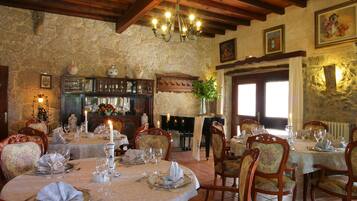A dining area with round tables set for a meal, wooden chairs, a chandelier, stone walls, and a fireplace.