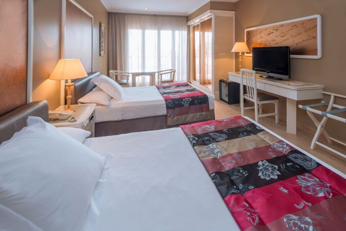 A hotel room with two beds, a desk, a television, and a window with curtains.