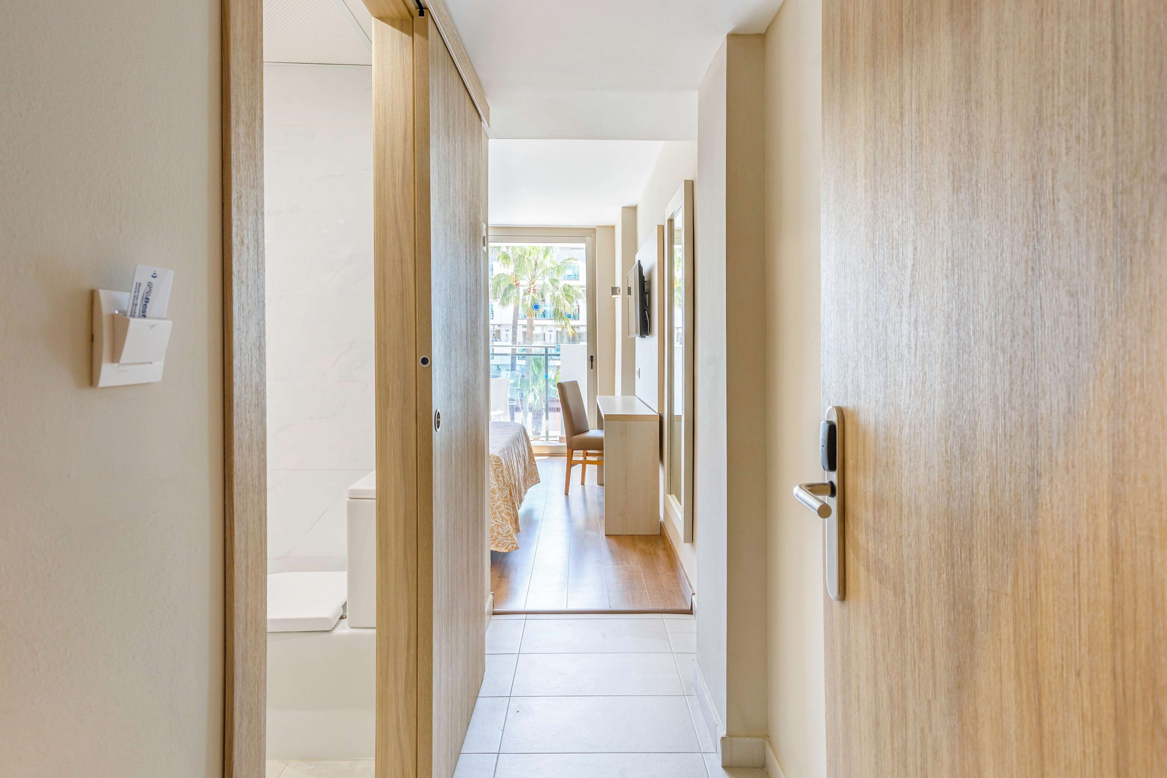 Un pasillo con puertas de madera, piso de baldosas blancas y vista a un comedor con mesa y sillas.