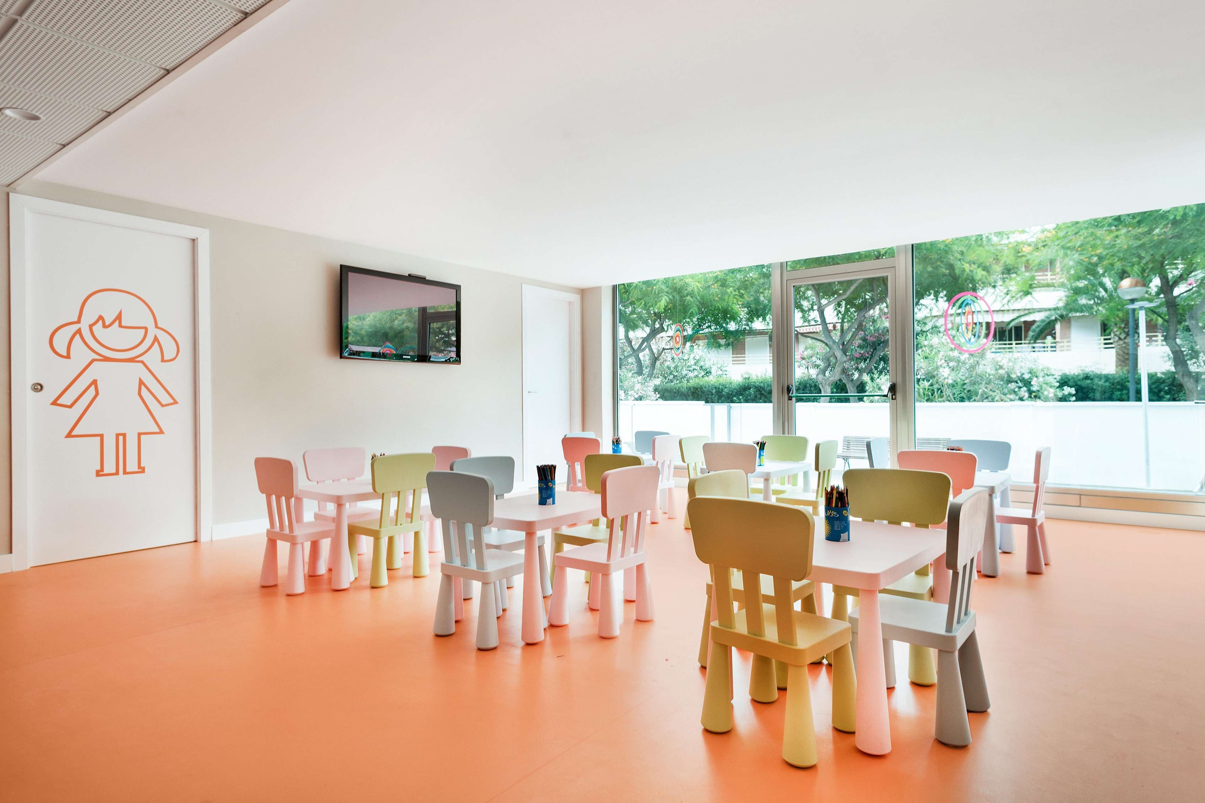 A brightly lit room with pastel-colored chairs and tables arranged in rows, large windows with a view of greenery outside.