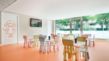 A brightly lit room with pastel-colored chairs and tables arranged in rows, large windows with a view of greenery outside.
