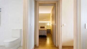 Un pasillo estrecho con un inodoro a la izquierda, una cama con mesita de noche y lámpara en el centro, y una puerta que da a otra habitación a la derecha.