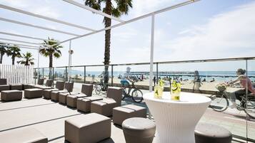 A rooftop lounge with outdoor seating, a table with drinks, and a view of the beach and ocean.