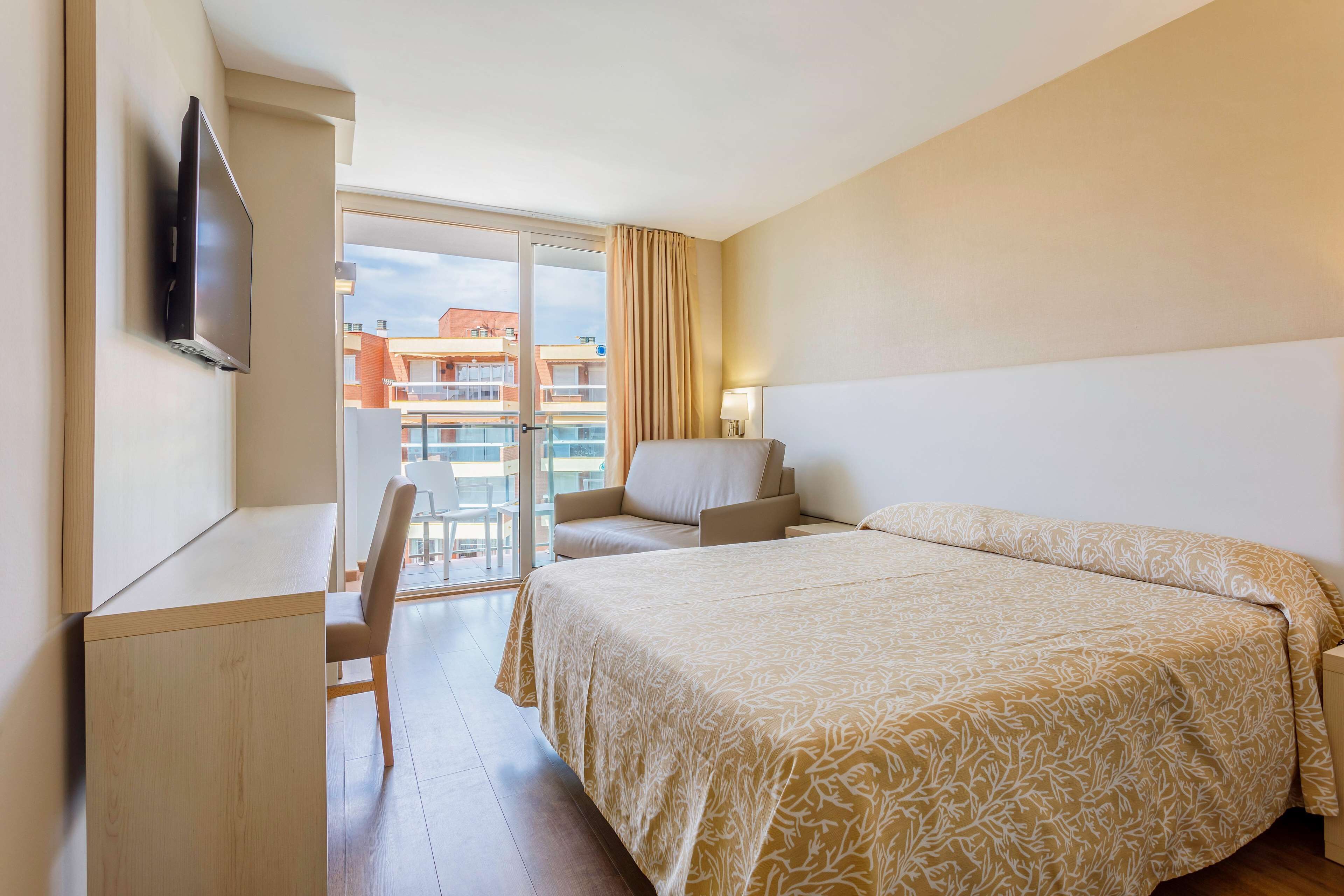 Habitación de hotel con cama, escritorio, silla, televisor y vista a edificios a través de un ventanal amplio.