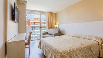 Habitación de hotel con cama, escritorio, silla, televisor y vista a edificios a través de un ventanal amplio.