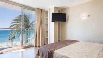Habitación de hotel con una cama grande, televisión montada en la pared y balcón con vista a la playa y palmeras.