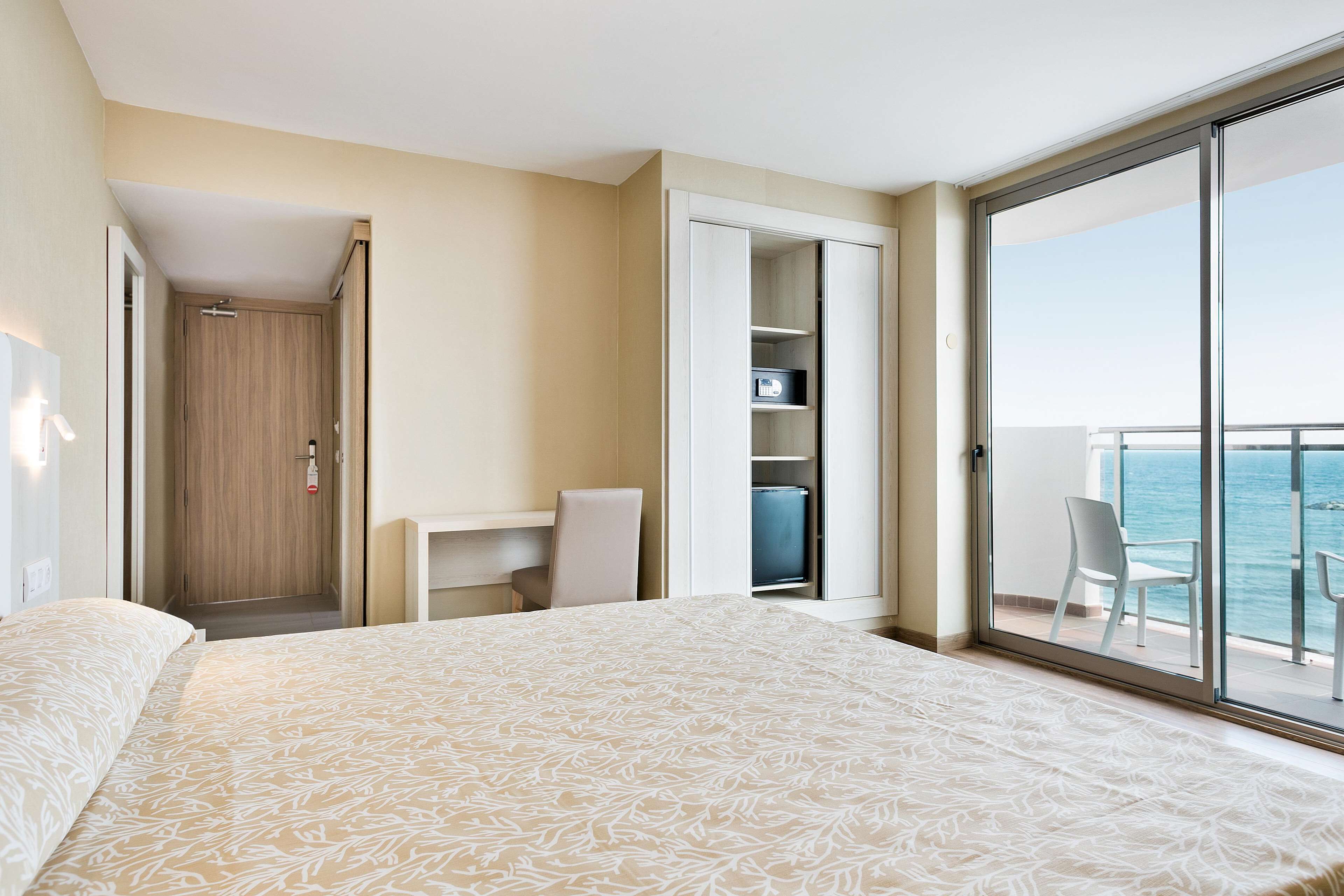 Habitación de hotel con cama, escritorio, silla, armario y balcón con vista al mar.