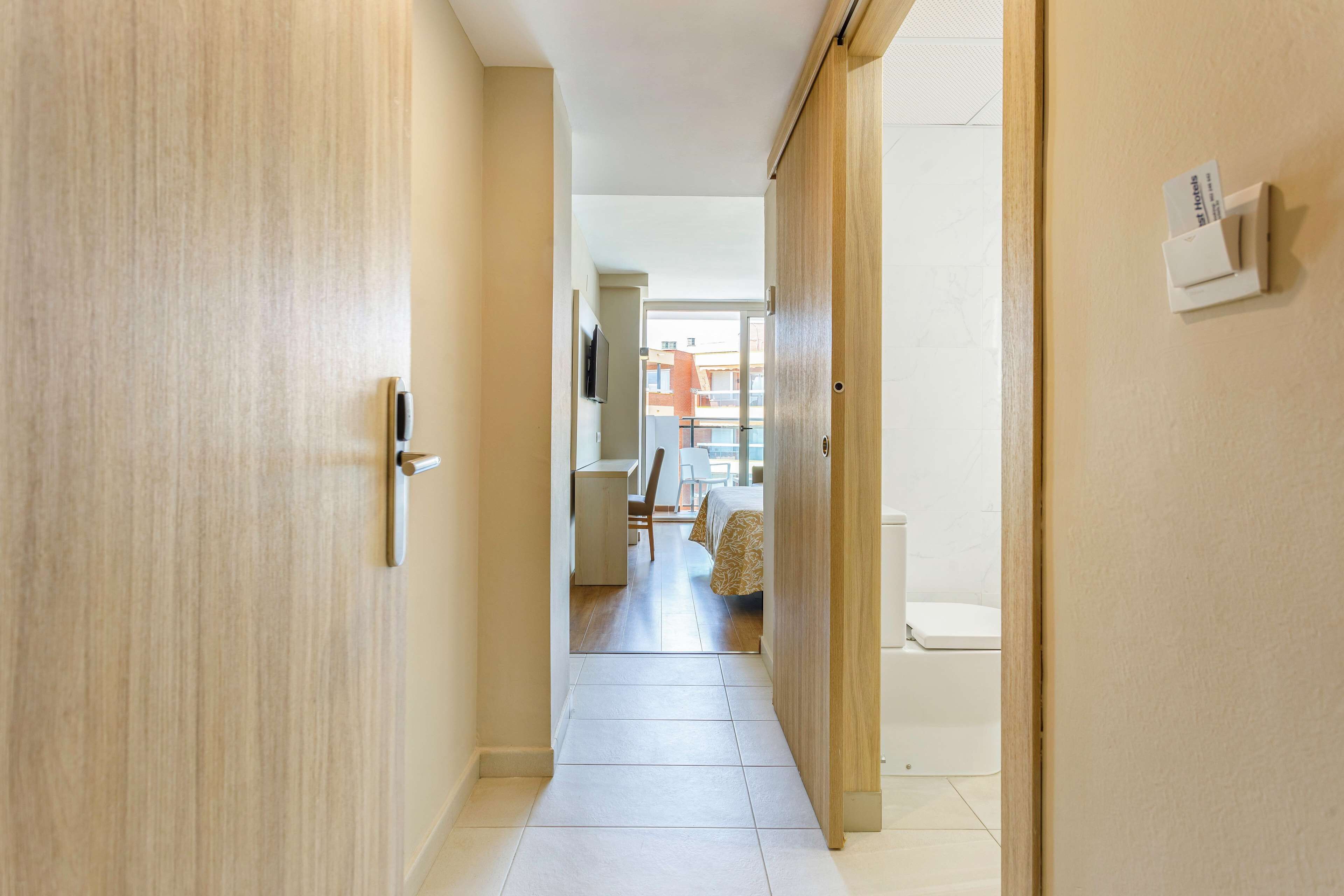 A hotel hallway with a wooden door, a bed, a desk, and a TV in the background.