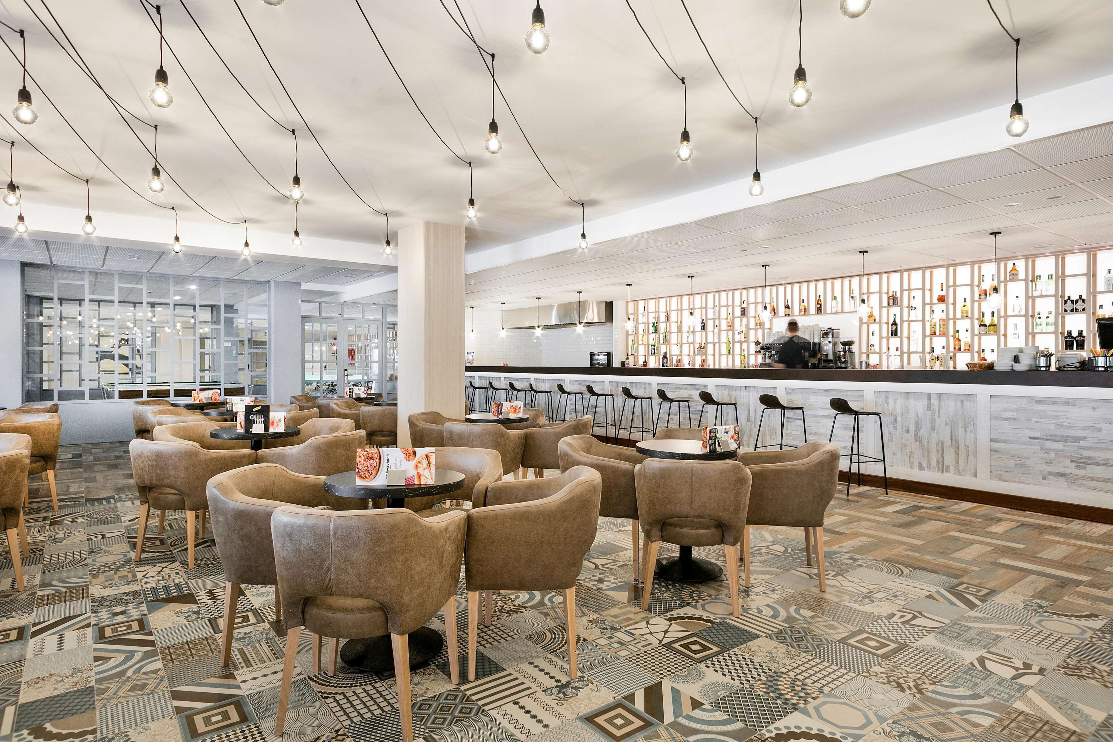 Zona de bar moderna con suelo estampado, diferentes distribuciones de asientos y una barra con gran variedad de botellas.