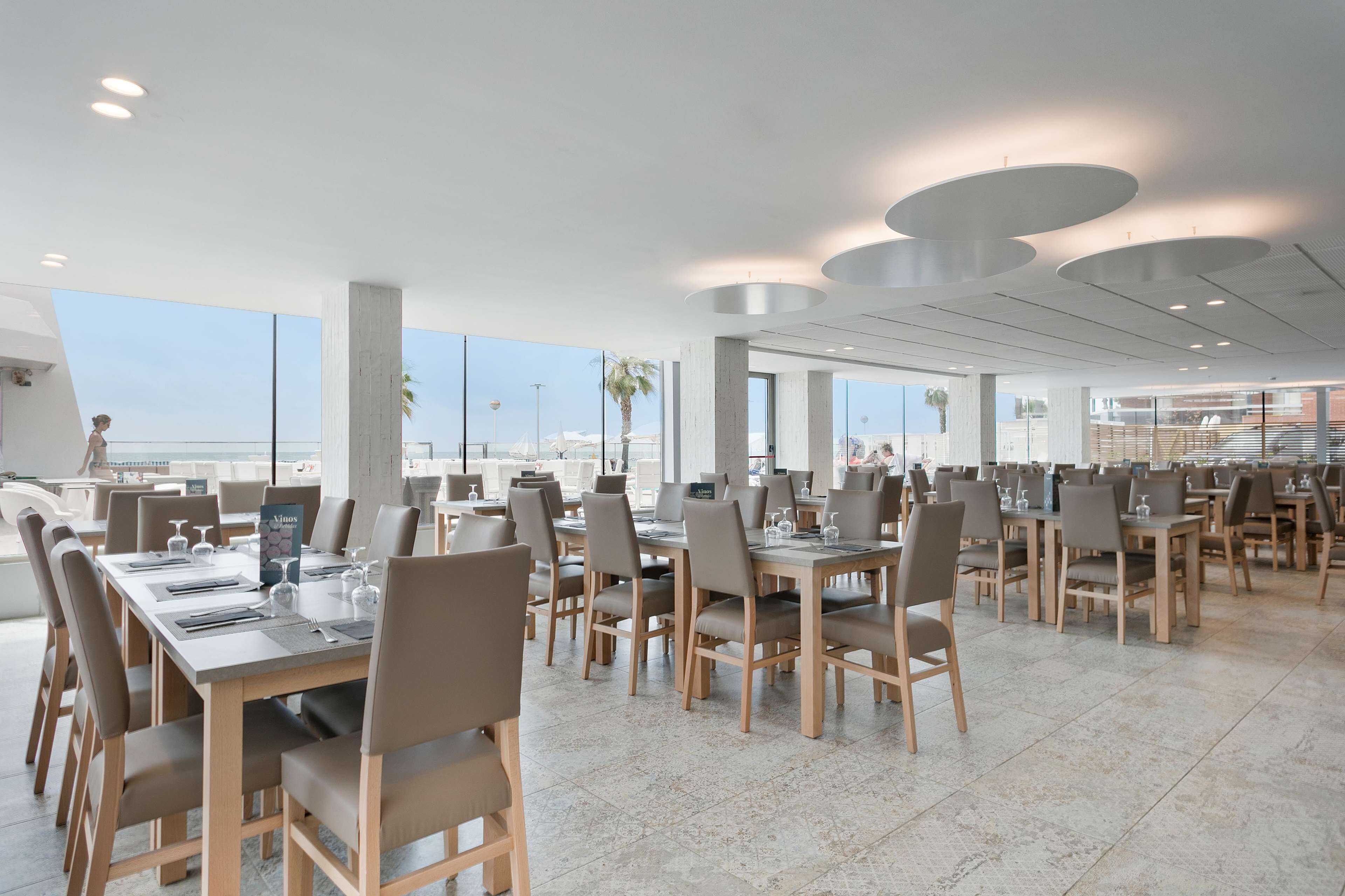 Un comedor amplio con varias mesas y sillas, ventanas grandes que ofrecen vista a la playa y un diseño de techo moderno.
