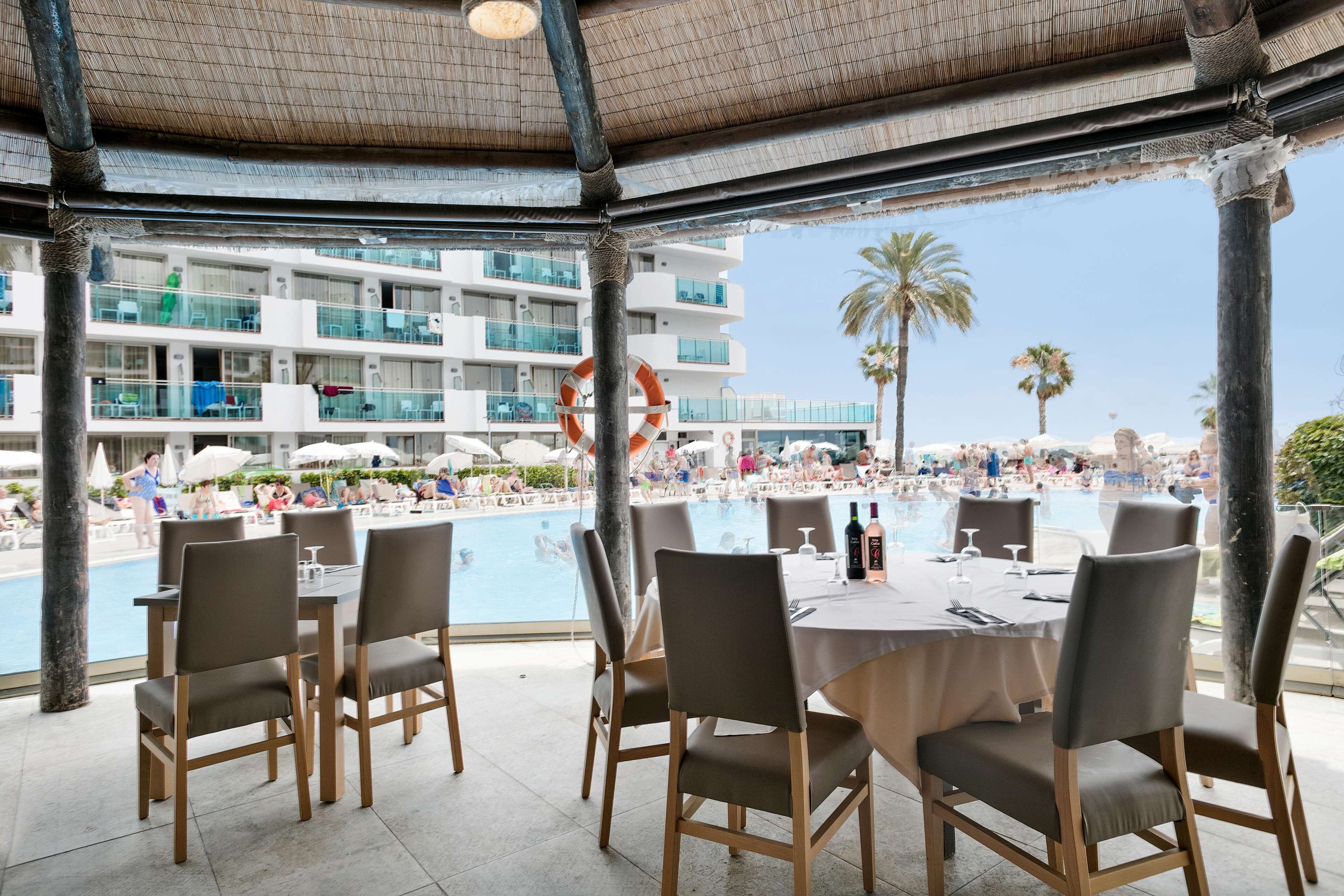 An outdoor dining area with a pool view, featuring wooden chairs and tables, a lifebuoy decoration, and a multi-story building in the background.