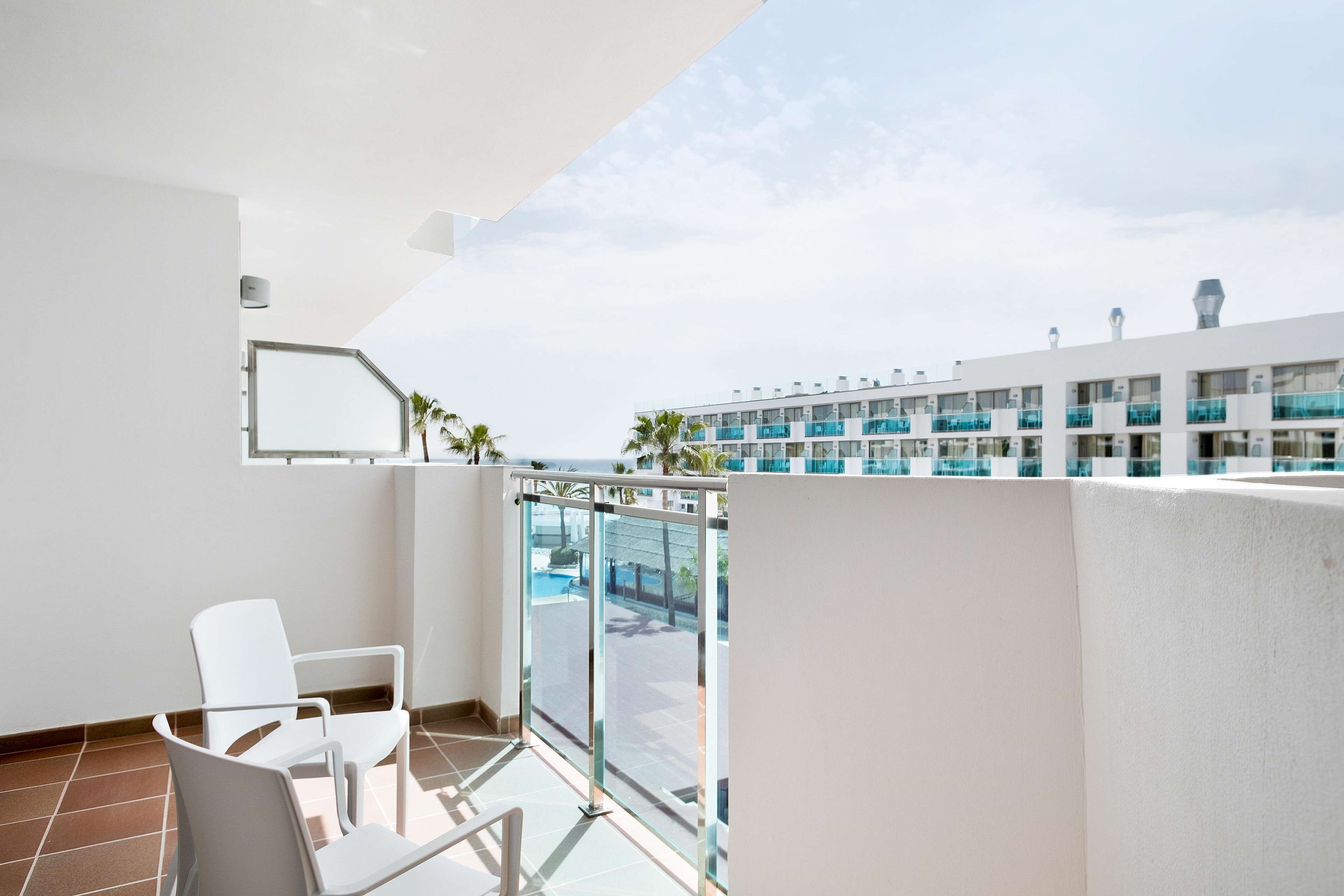 A balcony with white chairs, a glass door, and a view of a hotel building with multiple balconies.