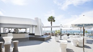 Un bar situé sur le toit d’un immeuble, avec des meubles blancs, un palmier et une enseigne indiquant « Meilleurs hôtels ».