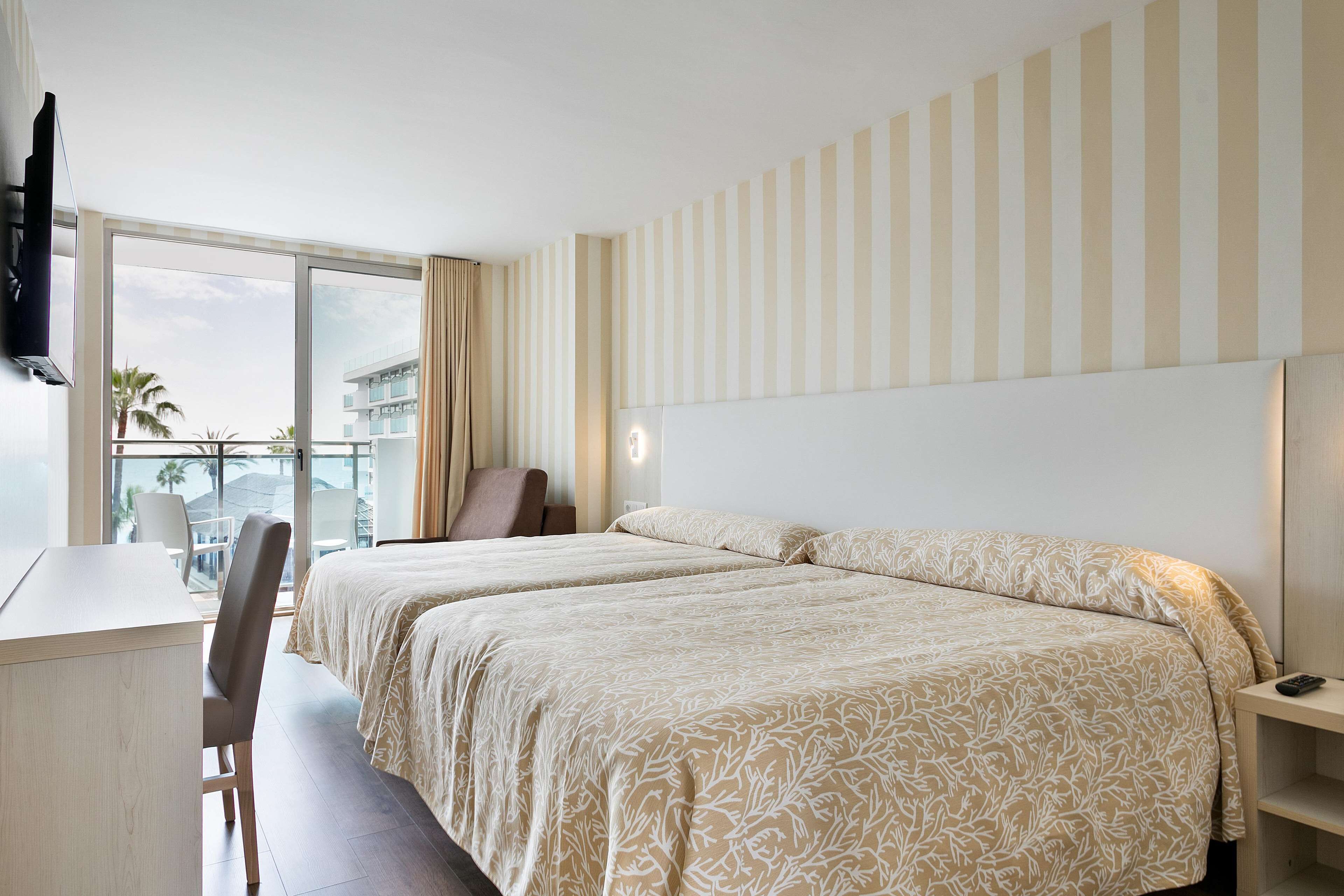 A hotel room with two beds, a desk, a chair, a TV, and a large window with a view of palm trees and buildings.