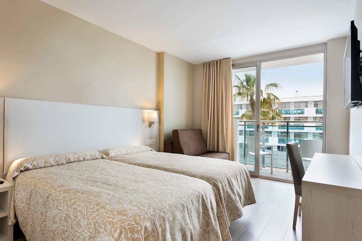 A hotel room with two beds, a chair, a desk, and a balcony with a view of palm trees and buildings.