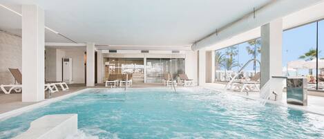 Una piscina interior con agua cristalina, escalones de cerámica blanca y sillas de descanso.