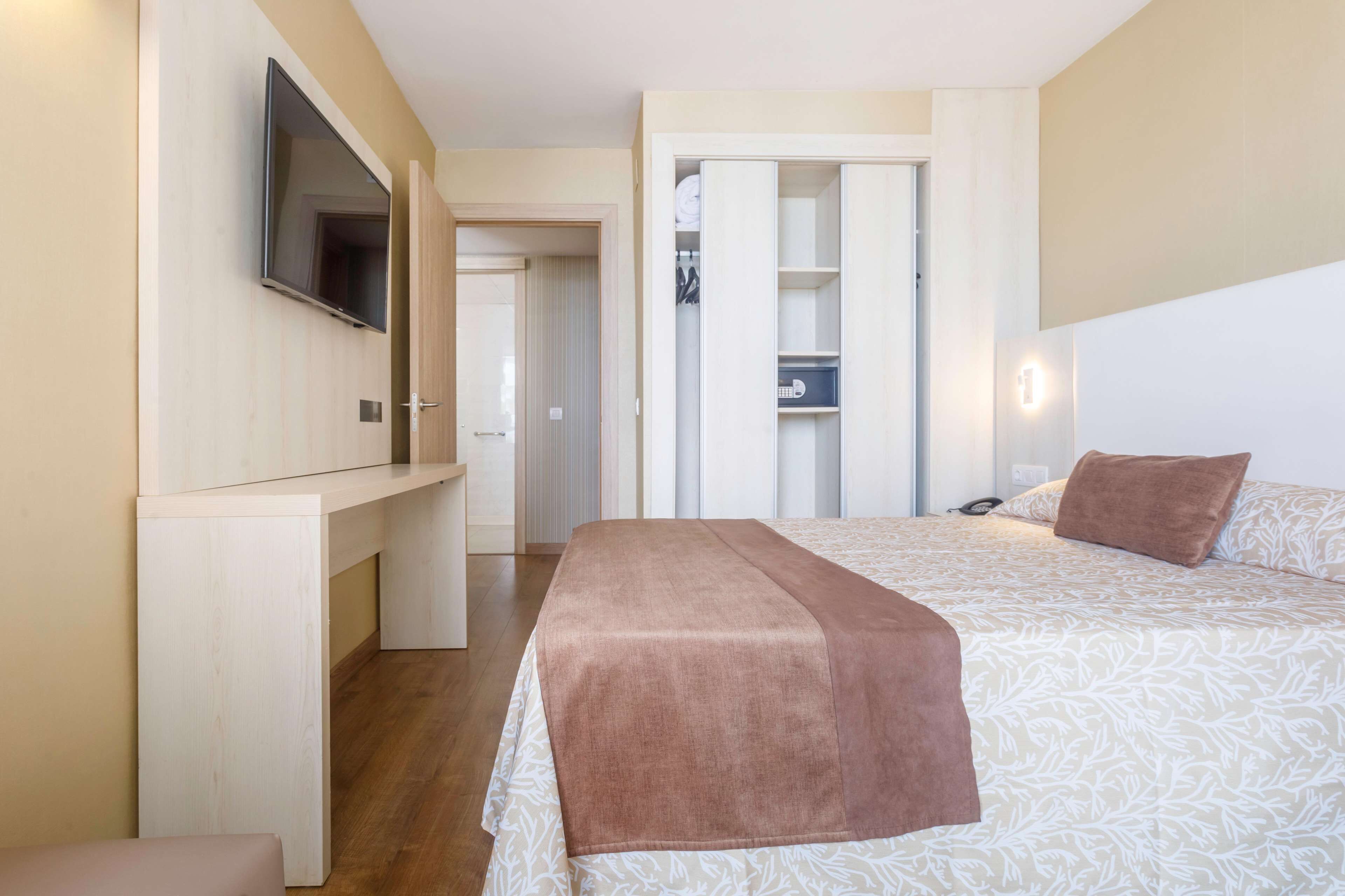 A bedroom with a bed, a TV mounted on the wall, a wardrobe, and a bedside table.