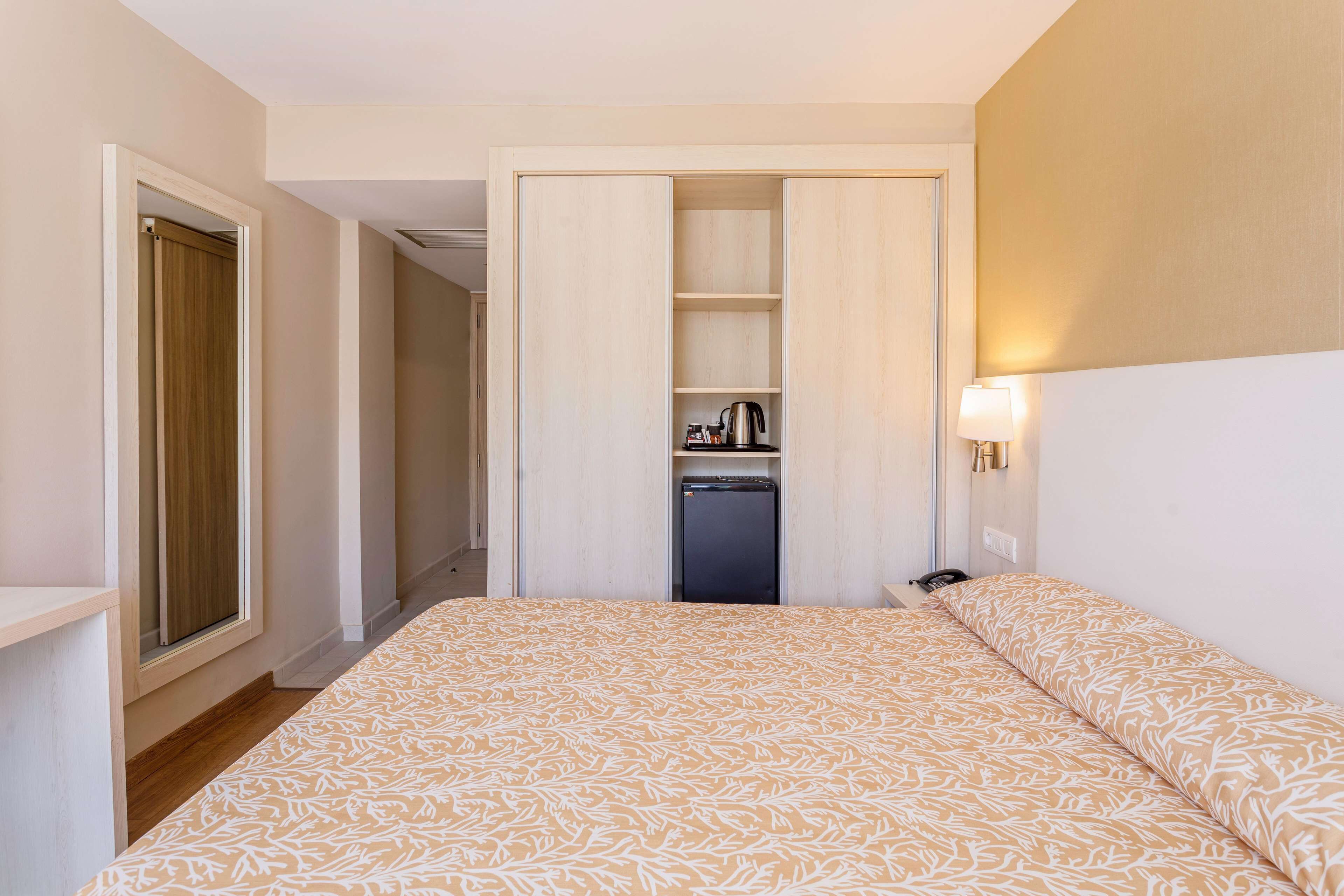 Habitació d'hotel amb un llit, un armari, una cuineta petita i una taula de nit amb una làmpada.