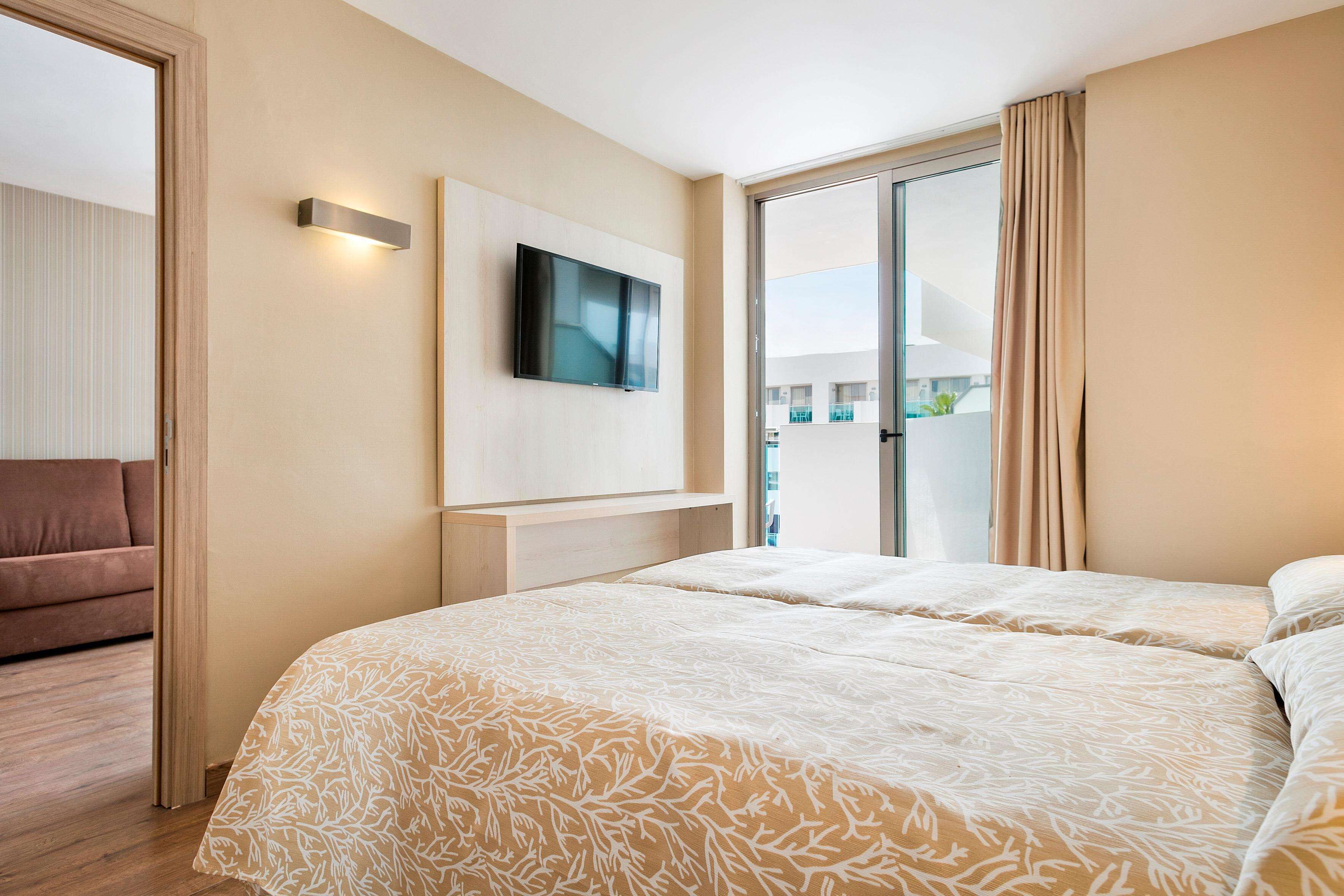 A hotel room with a large bed, a flat-screen TV mounted on the wall, and a sliding glass door leading to a balcony.