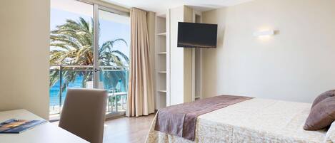 Habitación de hotel con cama, escritorio, silla, televisión y balcón con vista a palmeras y al mar.