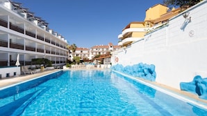 A swimming pool with a clear blue water surface, surrounded by a white building with balconies and a yellow building in the background. - Pez Azul (Puerto de la Cruz)
