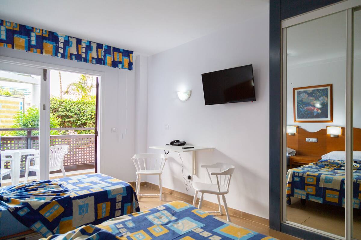 Une chambre d’hôtel avec un balcon, un bureau avec une chaise, un lit recouvert d’une courtepointe à motifs bleus et jaunes, un téléviseur fixé au mur et une fenêtre donnant sur un paysage verdoyant.
