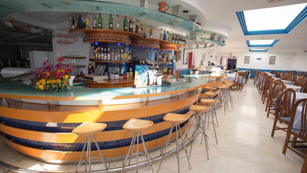 Een bar met krukken, een toonbank en diverse flessen. In de eetruimte staan tafels en stoelen.