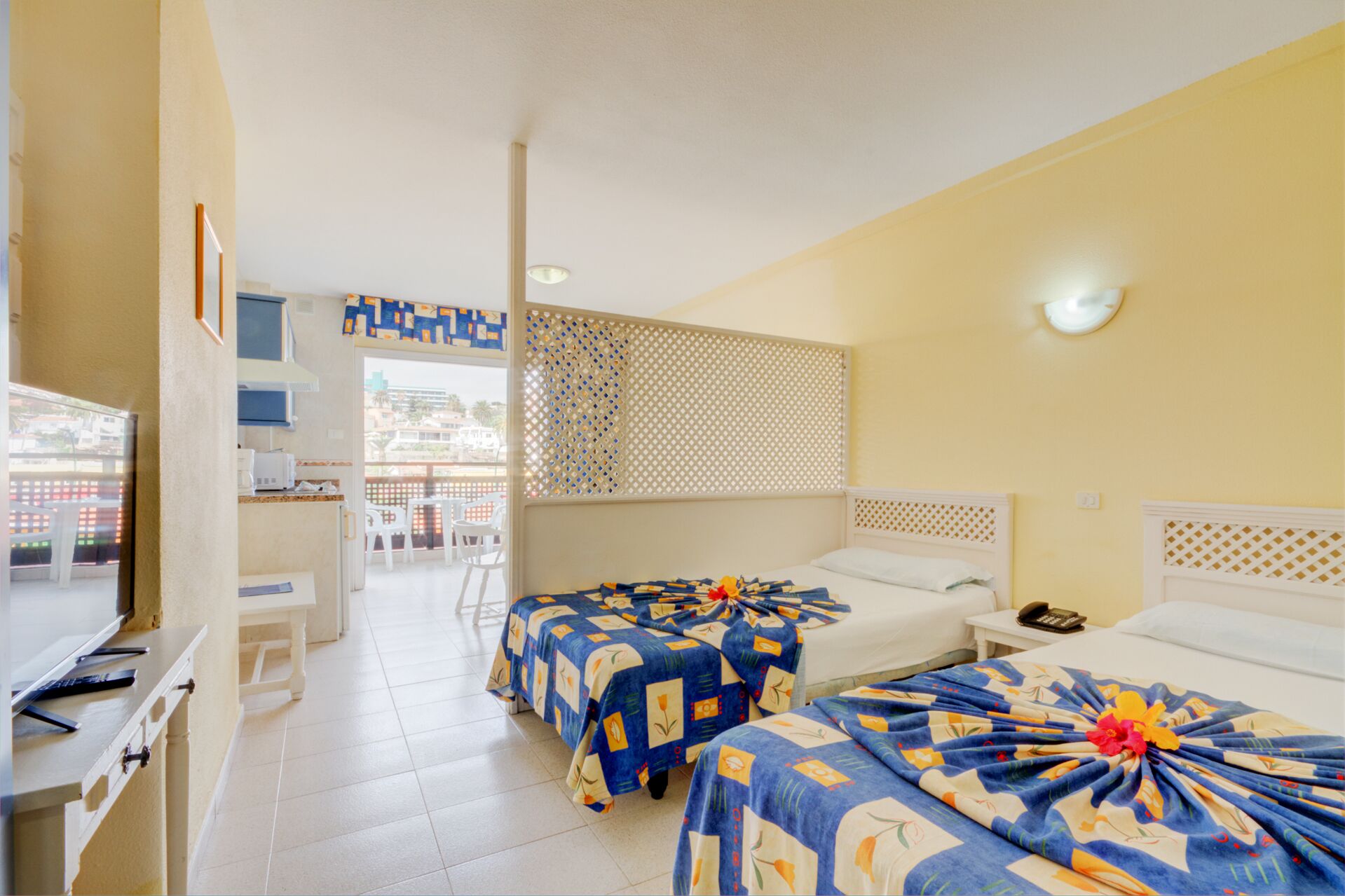 A hotel room with two beds, a balcony, a TV, and a kitchenette.