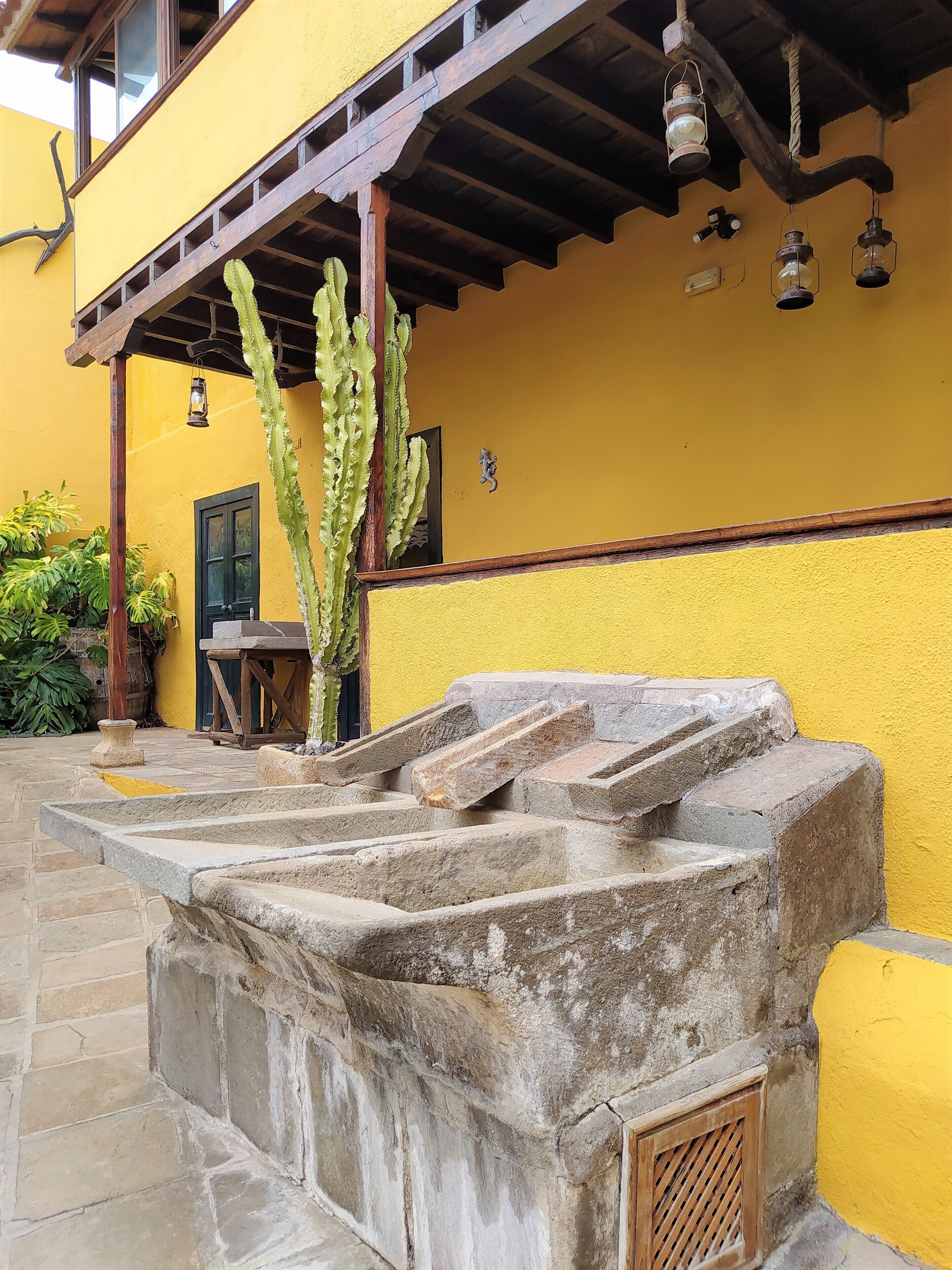 A stone water fountain with steps, a cactus, and a yellow building with wooden beams and lanterns.