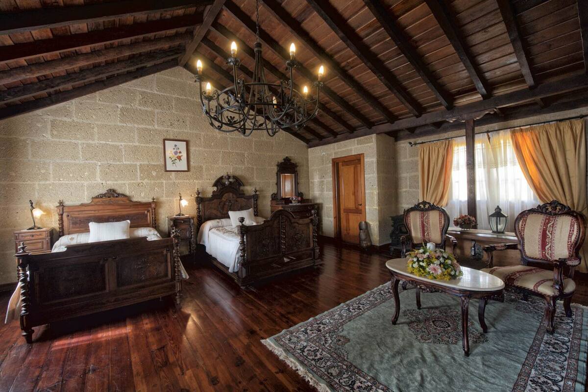 Une chambre traditionnelle avec un plafond en bois, un grand lit, un coin salon avec une table et des chaises, et un mur en pierre.