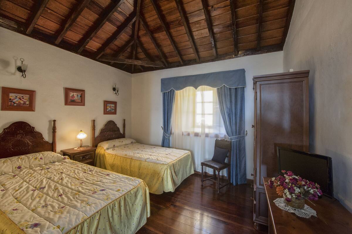 Une chambre avec deux lits, des têtes de lit en bois, une table de chevet, une chaise, une commode et une fenêtre avec des rideaux.