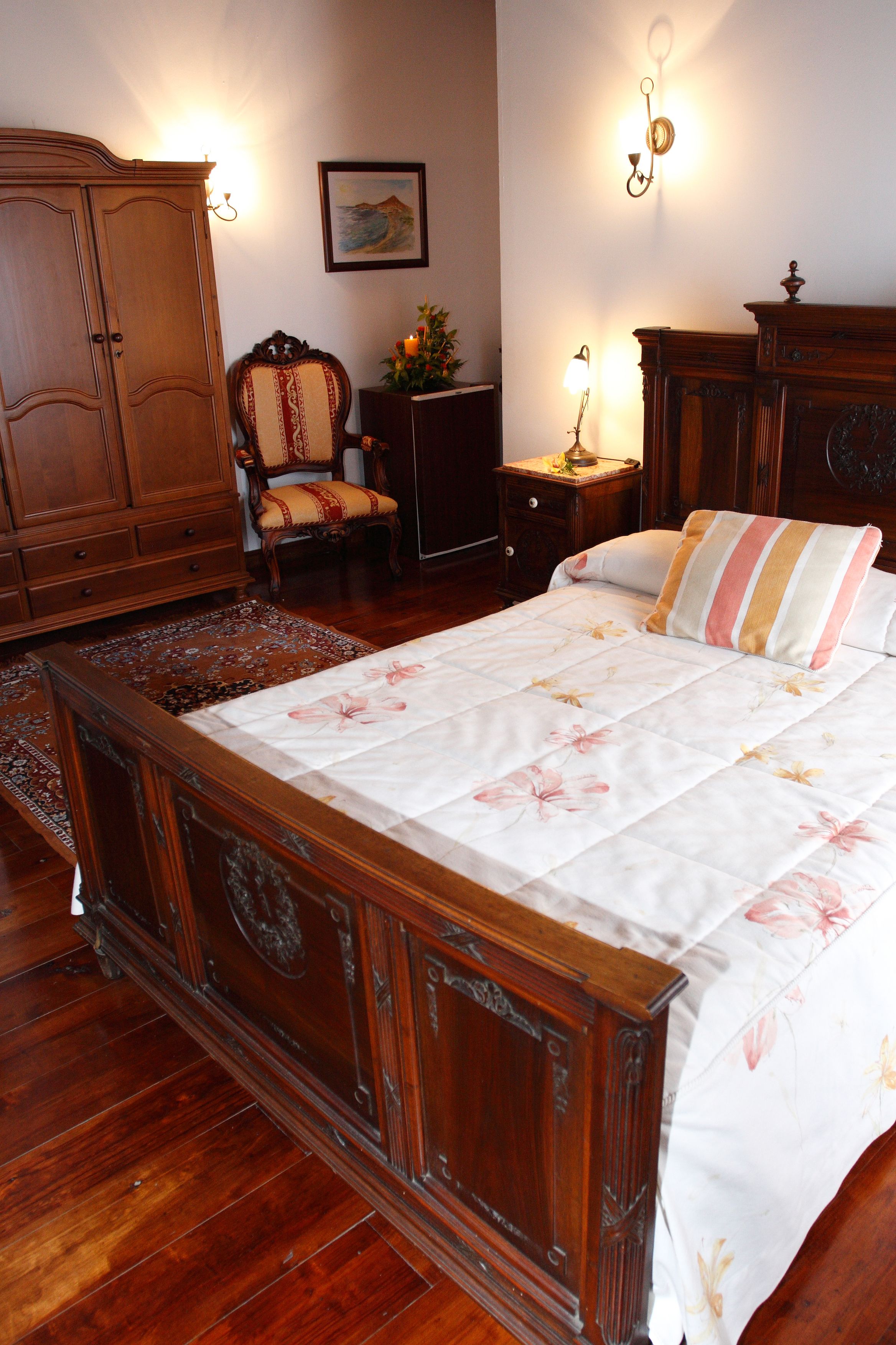 Un dormitorio con una cama de madera, un armario de madera, una silla, una mesita de noche y una lámpara de pared.