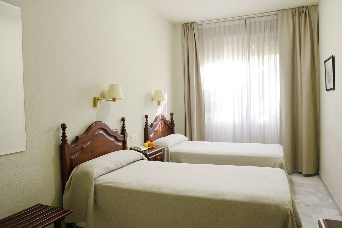 A hotel room with two single beds, a wooden nightstand with a lamp, a bench, and a large window with curtains.