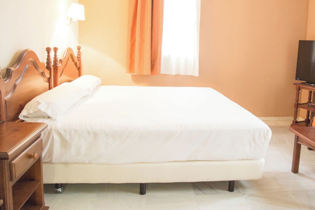 A hotel room with a bed, a wooden nightstand, a television, and an orange armchair.