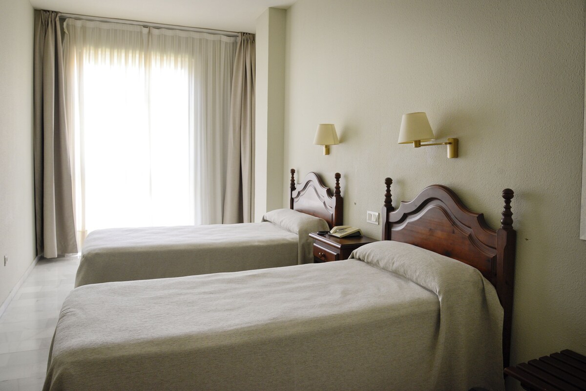 A hotel room with two beds, a nightstand, a lamp, and a window with curtains.