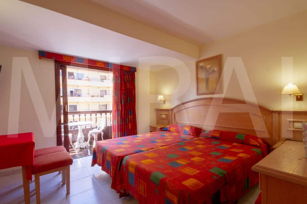 Habitación de hotel con dos camas, una silla roja, un balcón con vistas a edificios y una mesita de noche de madera con una lámpara.