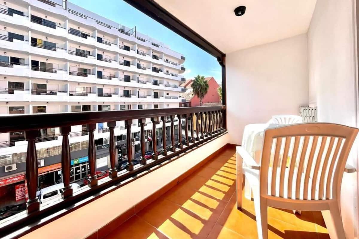 Un balcón con vista a un edificio de varios pisos, una silla de madera y una cama con cubrecama blanco.