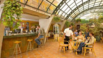 Cafetería interior con techo alto y paneles de vidrio, que incluye una zona de bar con taburetes y un área de mesas con sillas de mimbre.