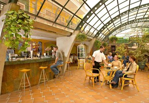 Cafetería interior con techo alto y paneles de vidrio, que incluye una zona de bar con taburetes y un área de mesas con sillas de mimbre.