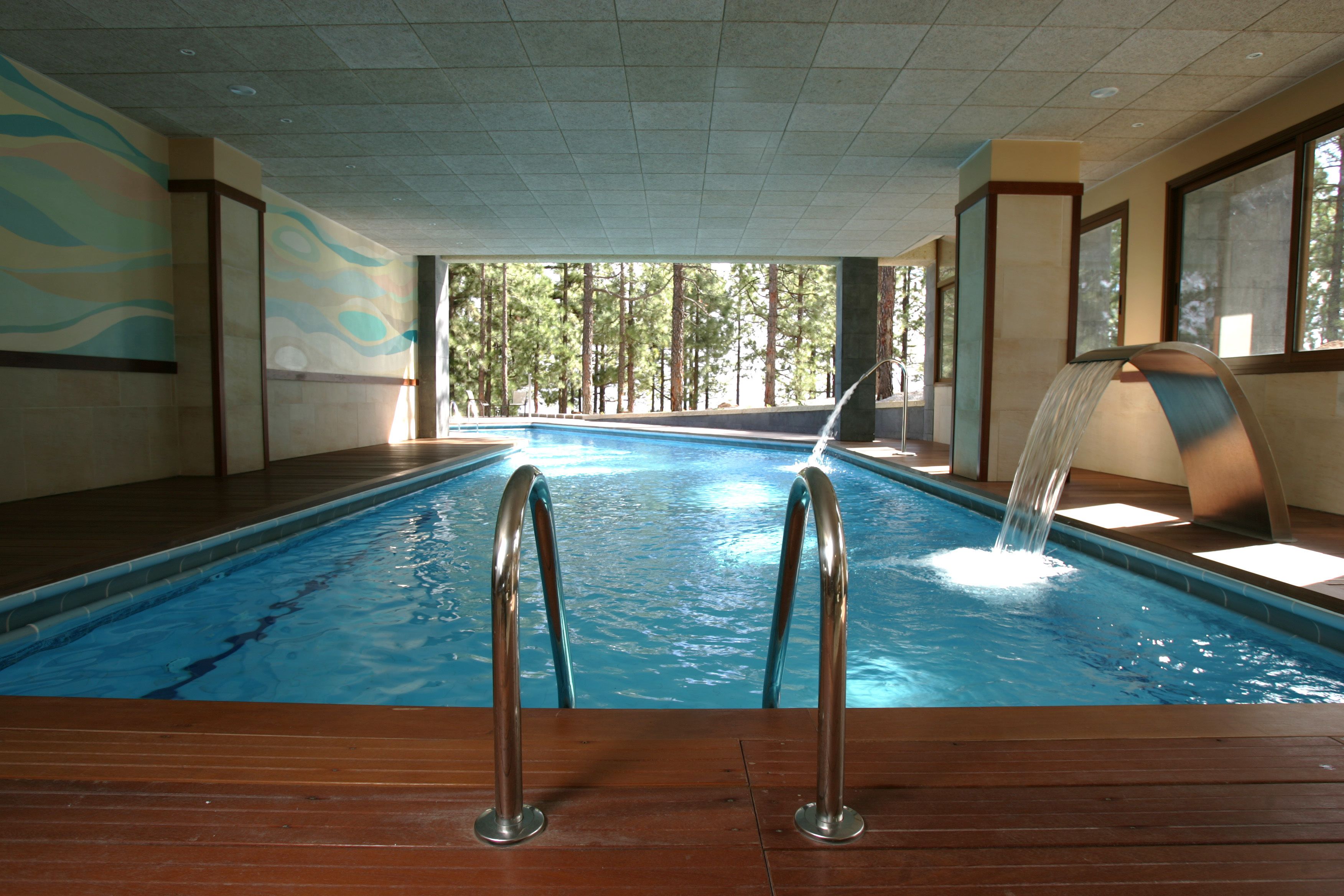 An indoor swimming pool with wooden flooring, metal railings, and a waterfall feature.