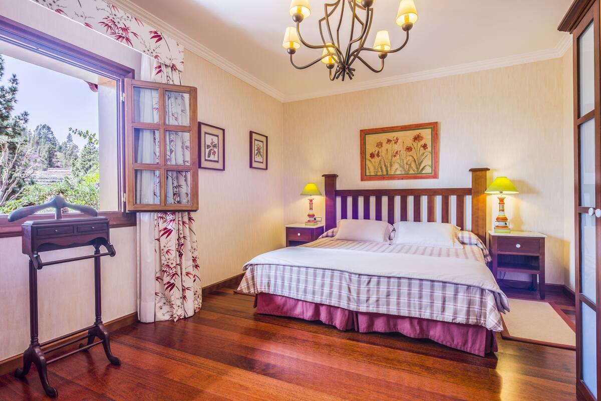A bedroom with a bed, two bedside tables, a chandelier, a window with curtains, and a desk with a chair.