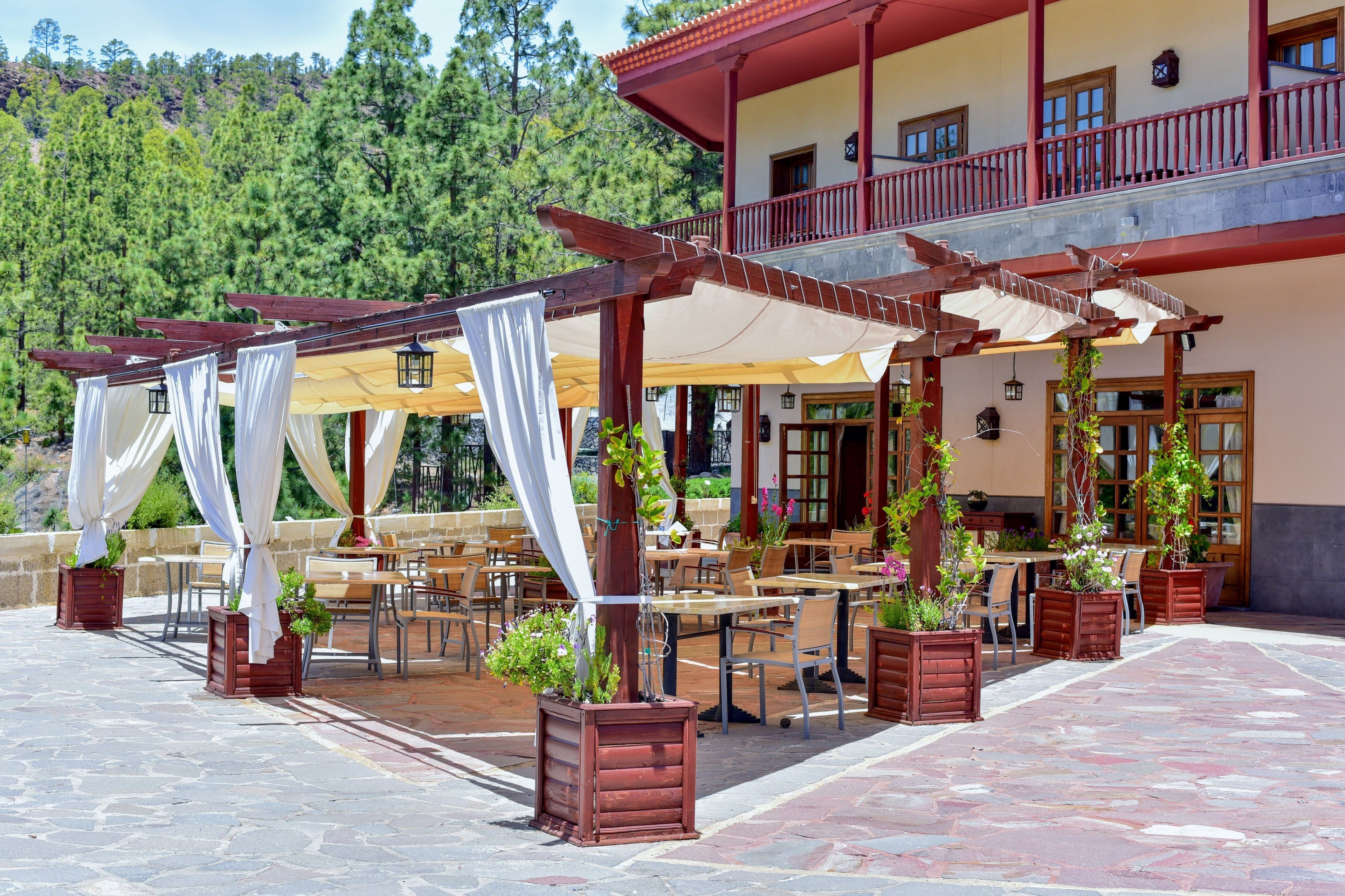 Un'area patio esterna con tavoli e sedie, circondata da piante in vaso e drappeggi bianchi.